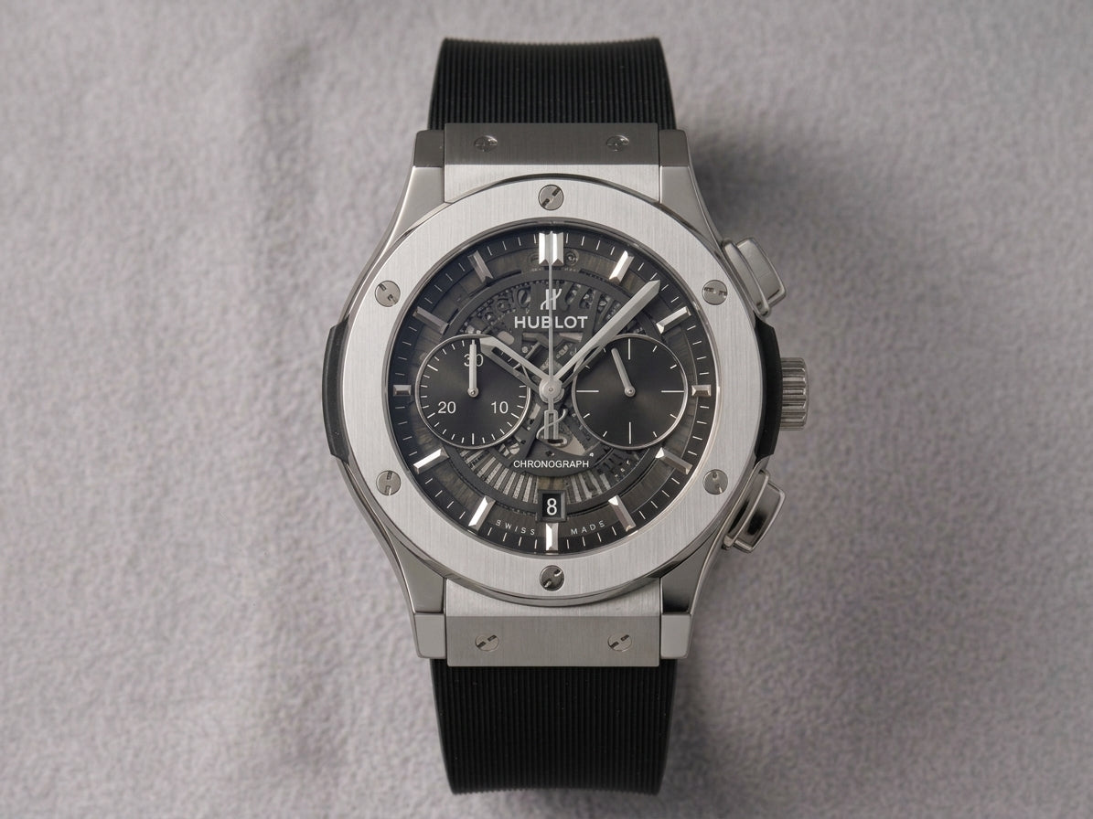 Hublot Big Bang chronograph watch with a black dial and black rubber strap, set in a silver-toned titanium case on a textured gray background.