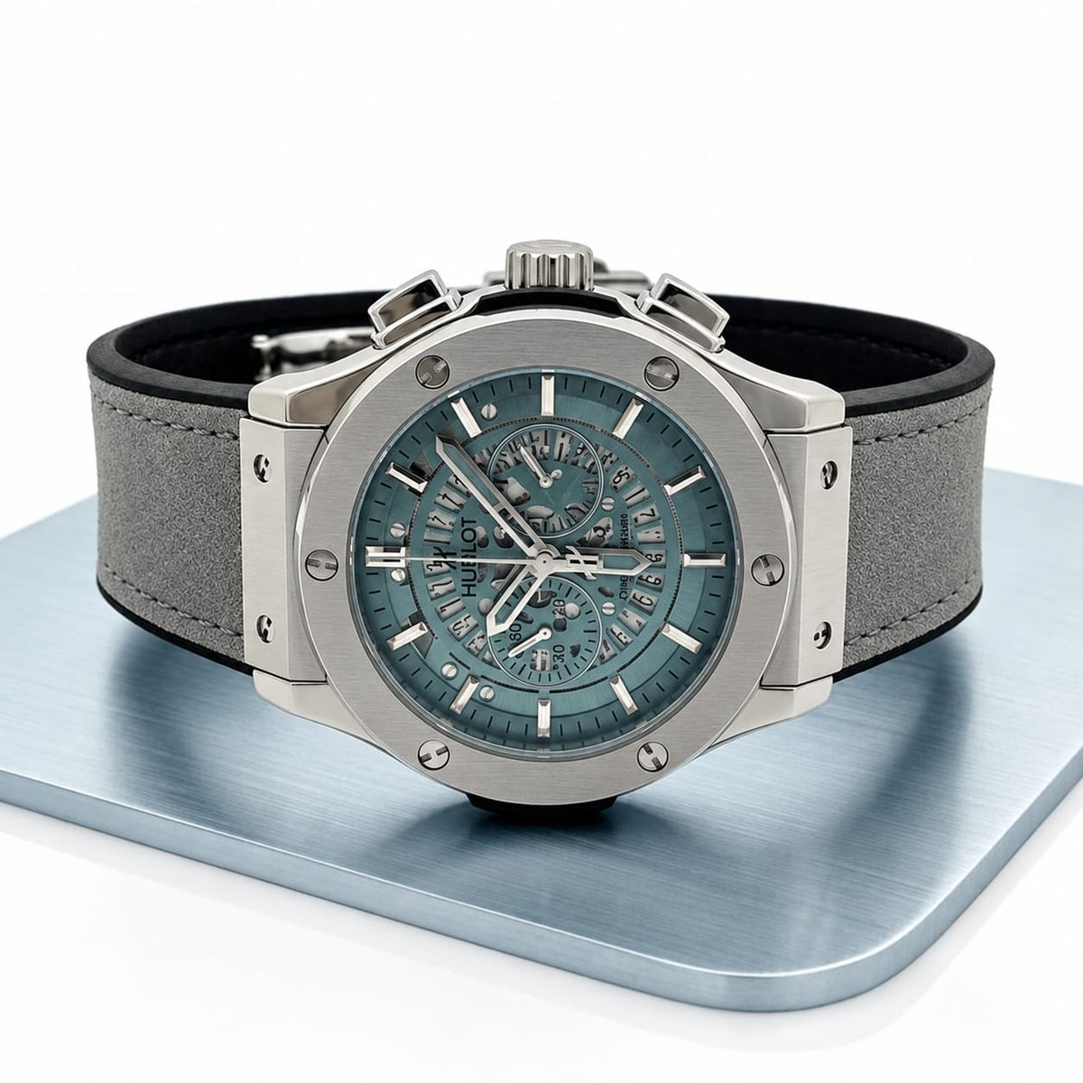 A silver-toned chronograph watch with a blue-green skeleton dial and a gray textured strap, displayed on a metallic blue-gray platform against a white background.